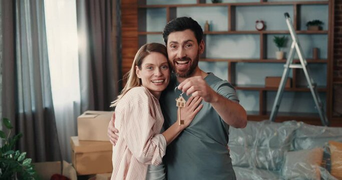 Portrait of loving  caucasian couple hugging while holding keys from new home, standing in spacious living room and looking at camera 
