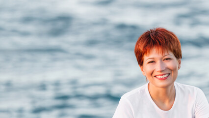 Fototapeta na wymiar Smiling woman with short bright red haircut. Caucasian woman looks happily on blue sea background. Horizontal banner with copy space.