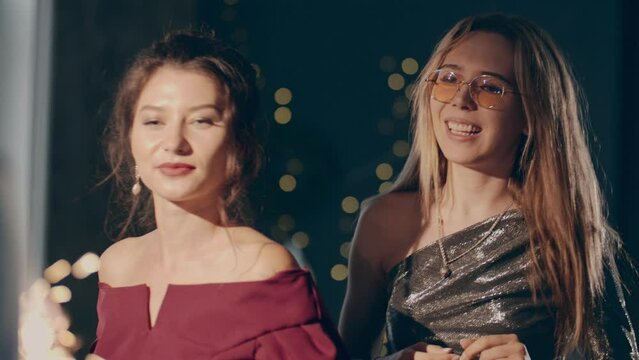 Women dancing with falling confetti and sparkling or glowing sparklers or fireworks. Close up couple of young multiracial girls wearing plush dresses enjoying glamorous party and having fun.