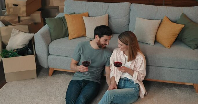 Couple in love holding glasses and drinking wine, having romatic mood and sitting on carpet at new apartment 