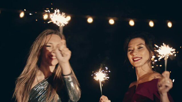 Two multiracial girlfriends in plush dresses dancing in dark at party holding and waving sparklers. Girls enjoying trendy or glamorous home party for Party Party, birthday. Party Party or Party.