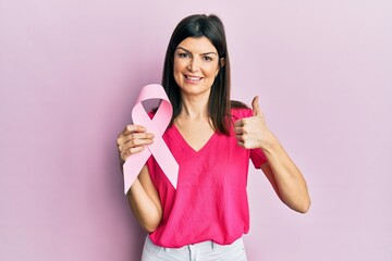 Young hispanic woman holding pink cancer ribbon smiling happy and positive, thumb up doing excellent and approval sign