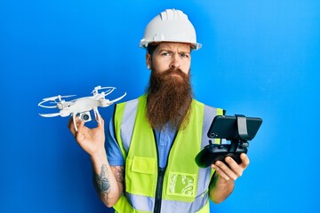 Redhead man with long beard wearing safety hard using drone with remote control skeptic and nervous, frowning upset because of problem. negative person.