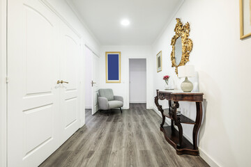 Distributor corridor of a house with eclectic decoration with white wood carpentry, vintage wood and marble locker, rococo gold-framed mirror and gray fabric armchair