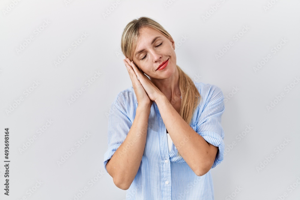 Sticker Beautiful blonde woman standing over white background sleeping tired dreaming and posing with hands together while smiling with closed eyes.