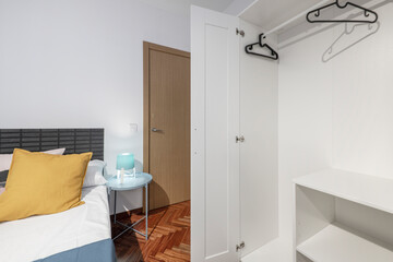 Open white wardrobe with shelves, coat rack and black hangers in bedroom with round blue nightstand and mahogany herringbone parquet flooring