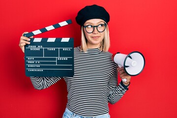 Beautiful blonde woman holding video film clapboard and megaphone smiling looking to the side and...