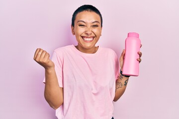 Beautiful hispanic woman with short hair holding shampoo bottle screaming proud, celebrating...