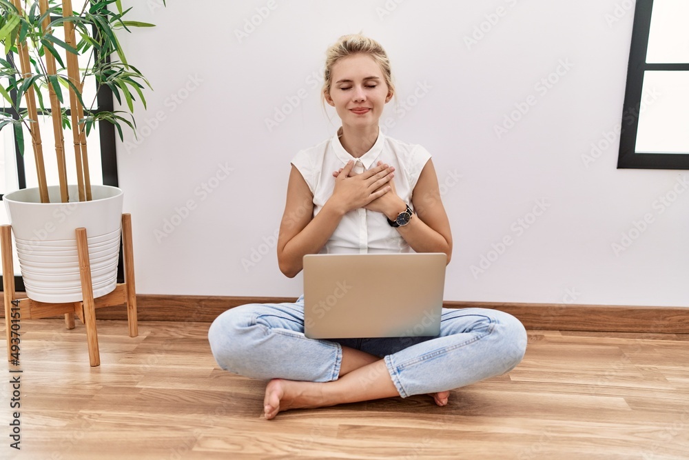 Canvas Prints Young blonde woman using computer laptop sitting on the floor at the living room smiling with hands on chest with closed eyes and grateful gesture on face. health concept.