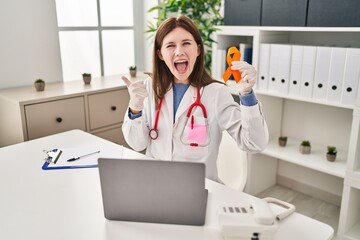 Young doctor woman holding awareness orange ribbon pointing thumb up to the side smiling happy with...