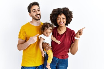 Interracial young family of black mother and hispanic father with daughter with a big smile on face, pointing with hand finger to the side looking at the camera.