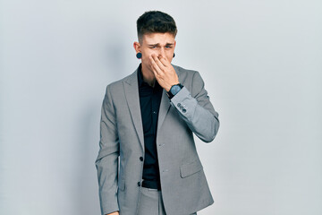Young caucasian boy with ears dilation wearing business jacket smelling something stinky and disgusting, intolerable smell, holding breath with fingers on nose. bad smell