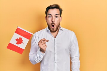 Handsome caucasian man with beard holding canada flag scared and amazed with open mouth for surprise, disbelief face