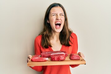 Young brunette woman holding board with raw meat angry and mad screaming frustrated and furious,...