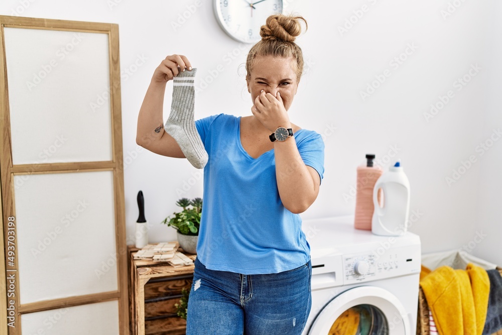 Wall mural Young caucasian woman holding dirty sock at laundry room smelling something stinky and disgusting, intolerable smell, holding breath with fingers on nose. bad smell