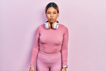 Beautiful hispanic woman wearing gym clothes and using headphones puffing cheeks with funny face. mouth inflated with air, crazy expression.