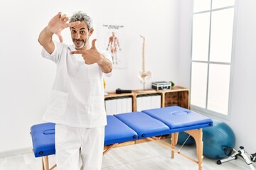 Obraz premium Middle age hispanic therapist man working at pain recovery clinic smiling making frame with hands and fingers with happy face. creativity and photography concept.