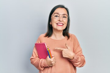 Middle age hispanic woman holding book and color pencils smiling happy pointing with hand and finger