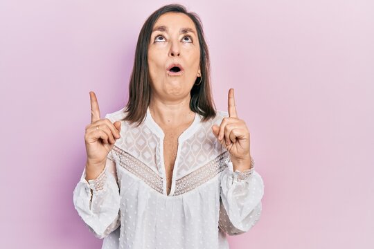 Middle age hispanic woman wearing casual clothes amazed and surprised looking up and pointing with fingers and raised arms.