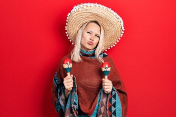 Beautiful caucasian blonde woman wearing festive mexican poncho and maracas looking at the camera blowing a kiss being lovely and sexy. love expression. - obrazy, fototapety, plakaty