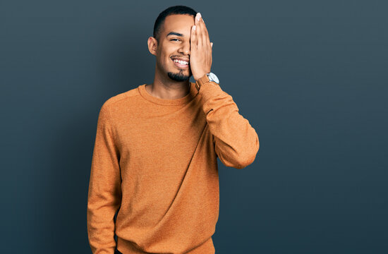 Young african american man wearing casual clothes covering one eye with hand, confident smile on face and surprise emotion.