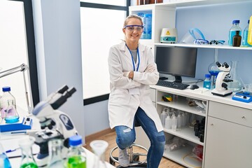 Young blonde woman wearing scientist uniform sitting with arms crossed gesture at laboratory
