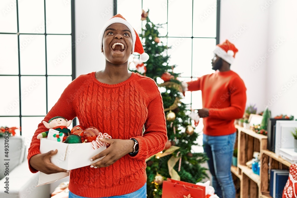 Poster young african american couple standing by christmas tree angry and mad screaming frustrated and furi