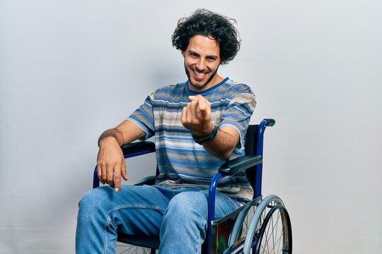 Handsome hispanic man sitting on wheelchair beckoning come here gesture with hand inviting welcoming happy and smiling