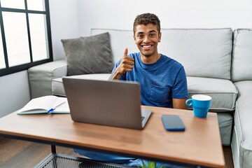 Young handsome hispanic man using laptop sitting on the floor doing happy thumbs up gesture with hand. approving expression looking at the camera showing success.