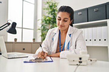 Young hispanic woman wearing doctor uniform writing medical report at clinic