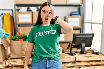 Young brunette woman wearing volunteer t shirt at donations stand doing ok gesture shocked with surprised face, eye looking through fingers. unbelieving expression.