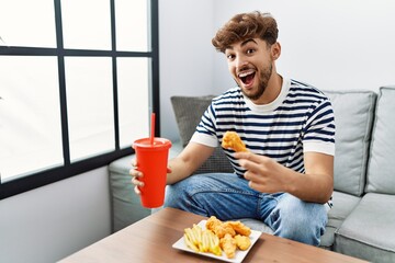 Young arab man smiling confident eating fried chicken drinking soda beverage at home