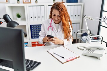 Obraz na płótnie Canvas Young redhead woman wearing doctor uniform using smartphone at hospital