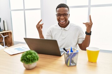 Young african man working at the office using computer laptop shouting with crazy expression doing rock symbol with hands up. music star. heavy music concept.