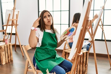 Young hispanic artist women painting on canvas at art studio shooting and killing oneself pointing hand and fingers to head like gun, suicide gesture.