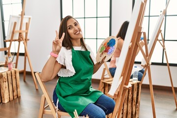 Young hispanic artist women painting on canvas at art studio smiling looking to the camera showing fingers doing victory sign. number two.