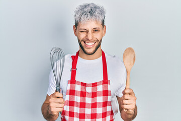 Young hispanic man with modern dyed hair wearing cook apron holding baker whisk and spoon winking...