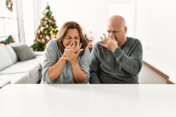 Middle age hispanic couple sitting on the table by christmas tree smelling something stinky and disgusting, intolerable smell, holding breath with fingers on nose. bad smell