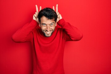 Handsome man with beard wearing casual red sweater posing funny and crazy with fingers on head as bunny ears, smiling cheerful