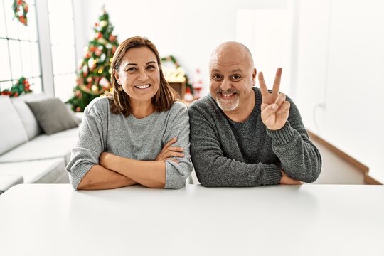Middle age hispanic couple sitting on the table by christmas tree showing and pointing up with fingers number two while smiling confident and happy.