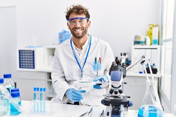 Young hispanic man wearing scientist uniform using loupe at laboratory