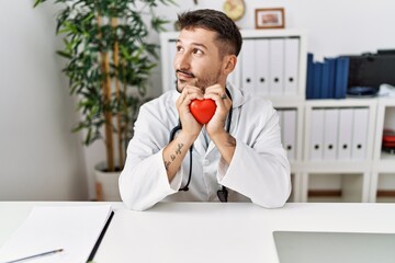 Young hispanic man wearing doctor uniform holding heart at clinic