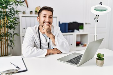 Young doctor working at the clinic using computer laptop looking confident at the camera smiling with crossed arms and hand raised on chin. thinking positive.