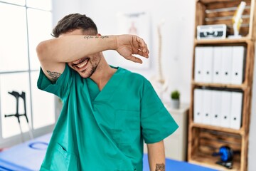 Young physiotherapist man working at pain recovery clinic covering eyes with arm smiling cheerful and funny. blind concept.