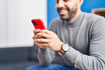 Young hispanic man using smartphone sitting on sofa at home
