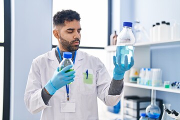 Young hispanic man wearing scientist uniform holding bottles at laboratory