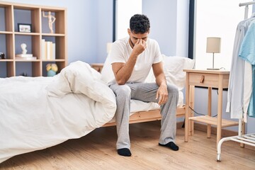 Young hispanic man suffering for headache sitting on bed at bedroom