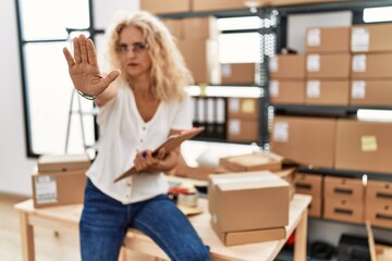 Middle age blonde woman working at small business ecommerce with open hand doing stop sign with serious and confident expression, defense gesture