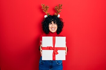 Young middle east woman wearing deer christmas hat holding gift smiling and laughing hard out loud because funny crazy joke.