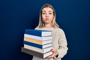 Beautiful caucasian woman holding a pile of books puffing cheeks with funny face. mouth inflated with air, catching air.
